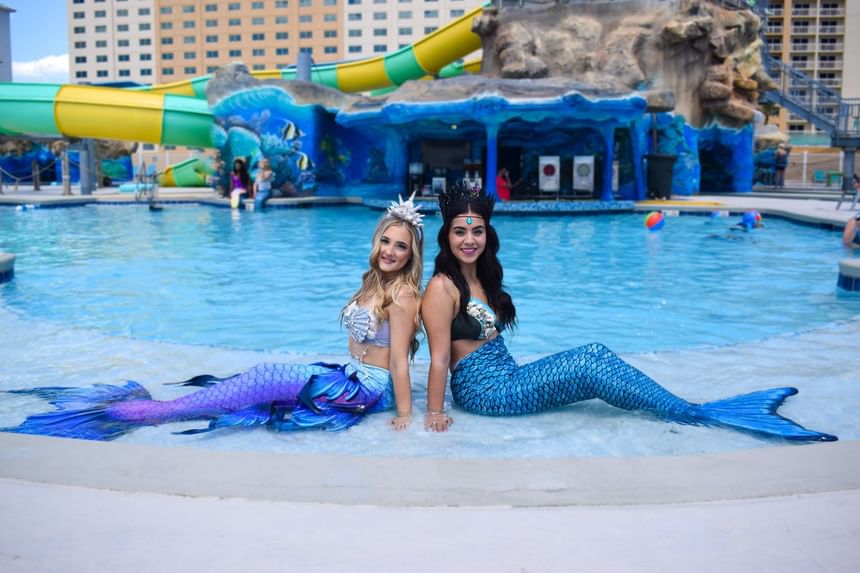 Two professional mermaids posing by the edge of a vibrant tropical swimming pool at Margaritaville Resort Biloxi