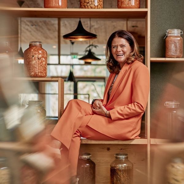 Eine Frau in einem orangefarbenen Anzug sitzt auf einem Regal mit Gläsern und lacht.