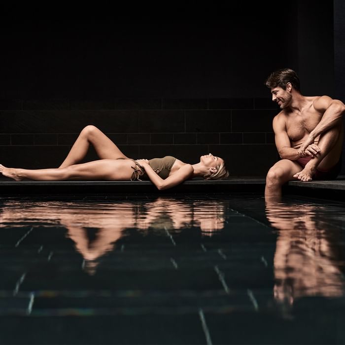 A man and a woman in swimsuits sit by a pool, gazing at each other.