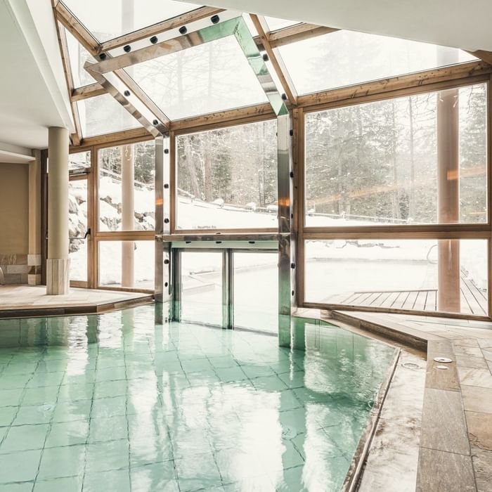Piscina interna con tetto in vetro, circondata da finestre grandi che mostrano una vista esterna innevata.