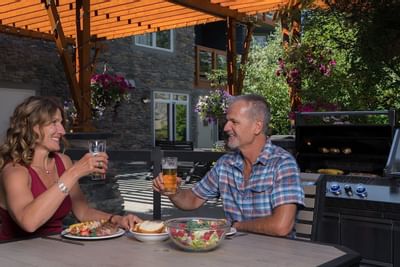 Couple enjoying a meal and drinks outdoors with a barbecue grill in the background at Blackstone Mountain Lodge