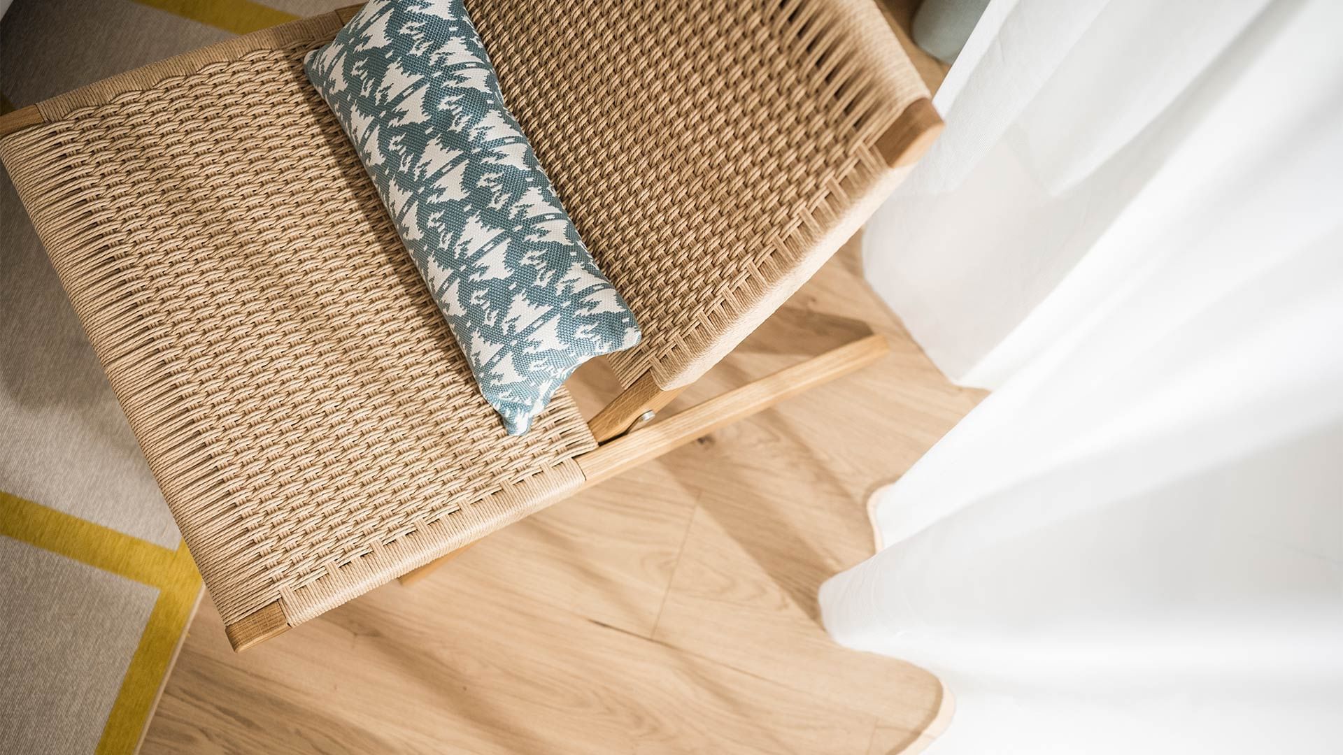 Woven chair with a blue and white pillow on a wooden floor next to a white curtain.