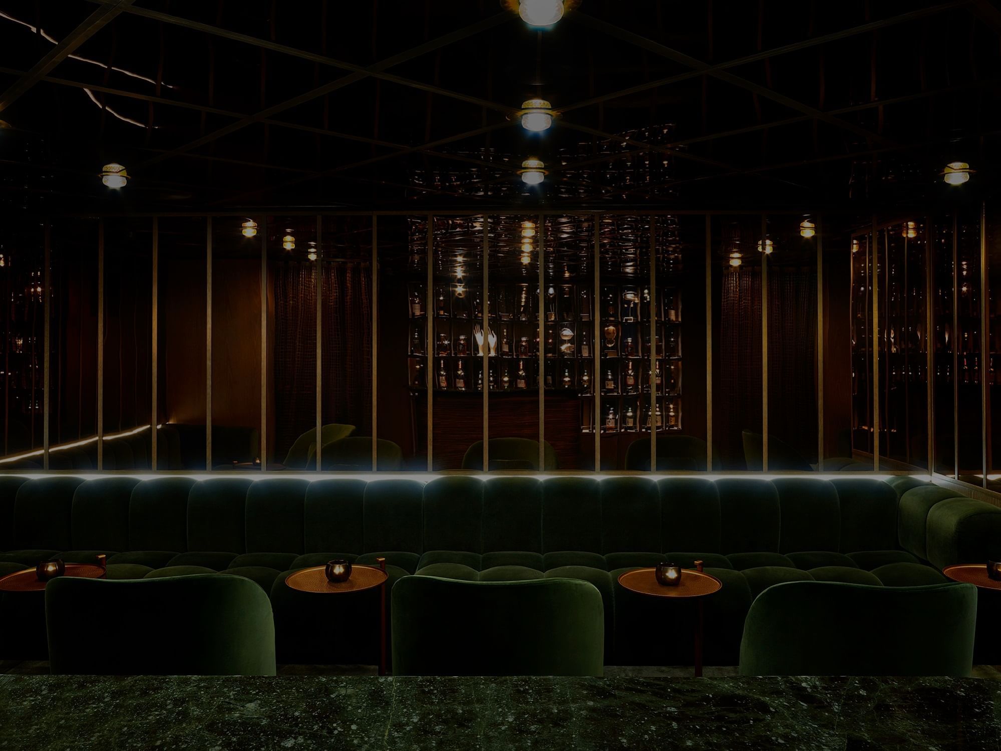 A dimly lit lounge featuring plush green seating, wooden tables, and shelves lined with liquor bottles at The Londoner Hotel