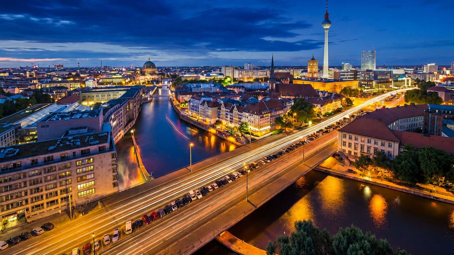 Aerial view of Berlin's illuminated skyline at dusk featuring the TV Tower and Spree River with light trails from traffic.