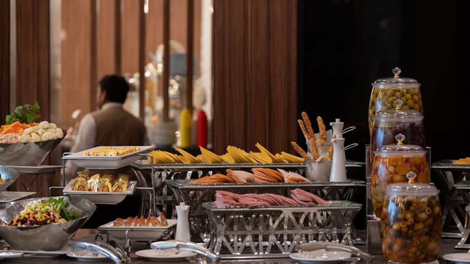 Cold cuts and cheese slices on silver stands by jars of olives at a breakfast buffet at Warwick Al Jubail