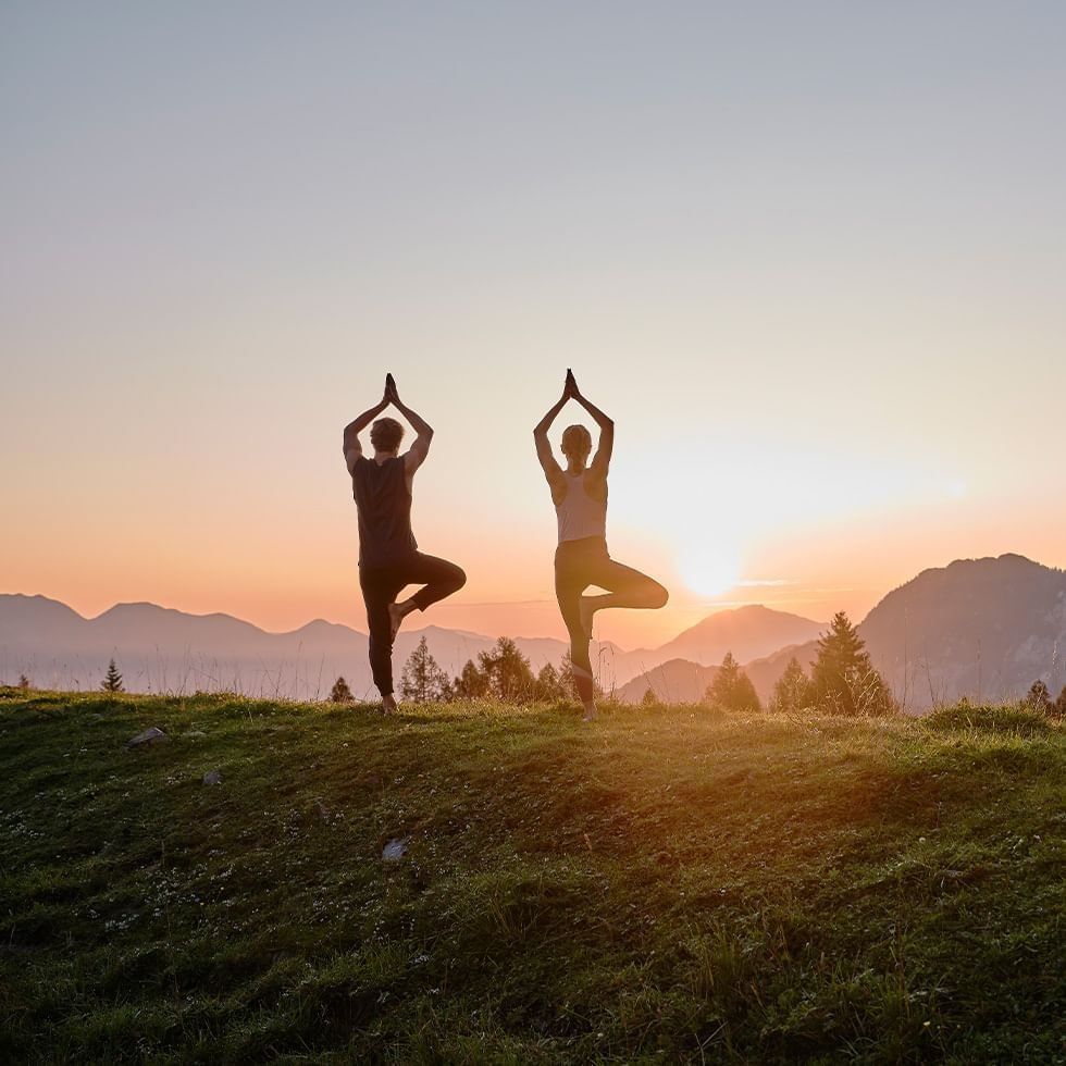 Two people performing yoga on a grassy hill at sunset with mountains in the background.