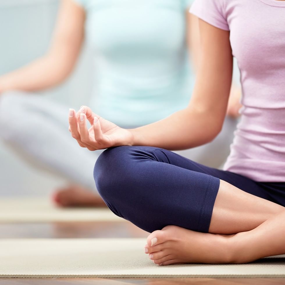 Zwei Frauen meditieren im Lotussitz auf Yogamatten, eine im Vordergrund, die andere im Hintergrund.