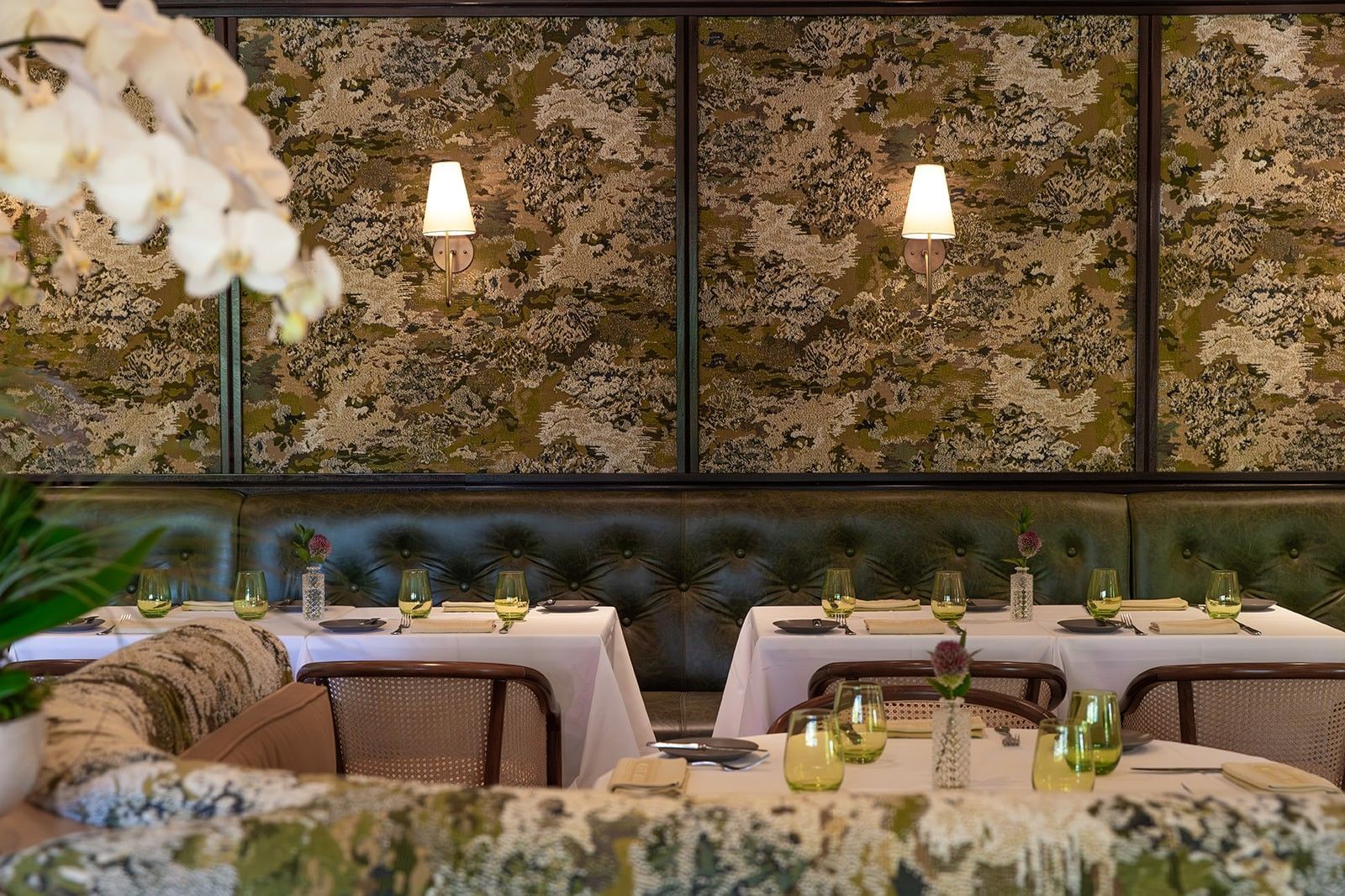 A dining area with decorative walls, green upholstered seating, and tables set with glassware and flowers.