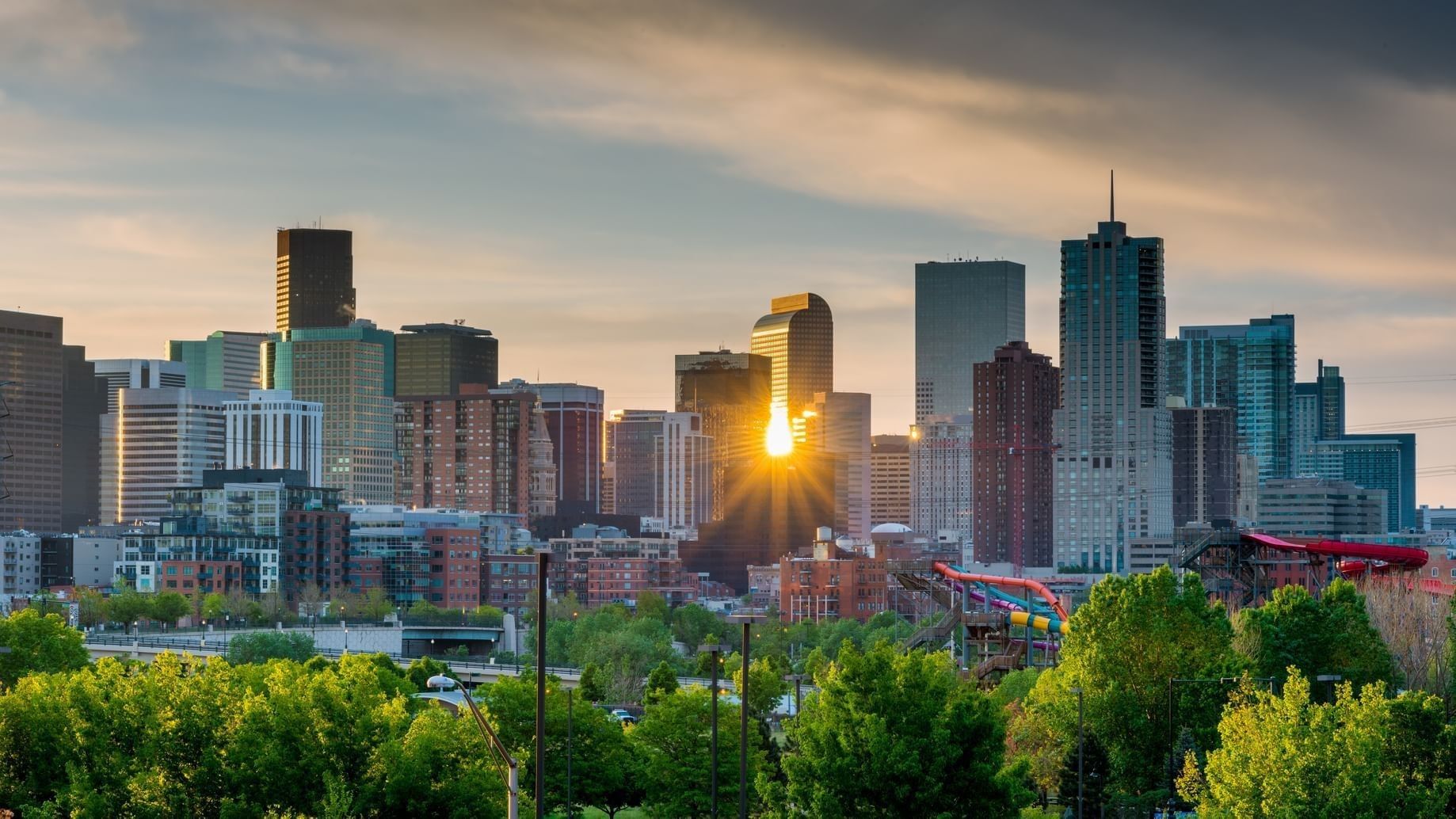 A cityscape at sunset with skyscrapers and trees in the foreground.