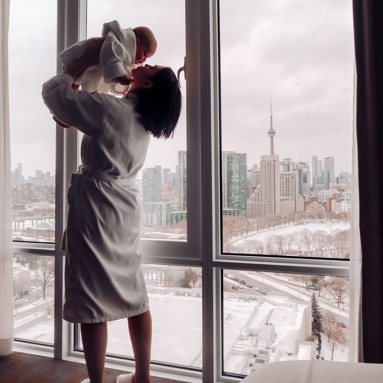 Woman in robe holding baby facing high rise window with snowy Toronto skyline and CN Tower view at Hotel X Toronto