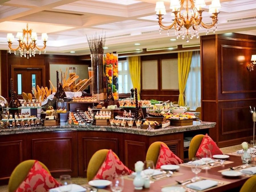 Ornate dessert buffet island by dining tables under a gold chandelier in a wood-paneled room at Warwick Doha Hotel