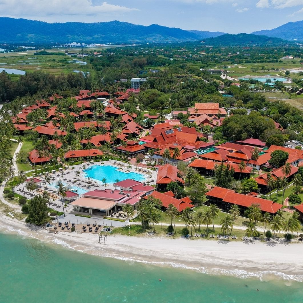 Aerial view of a resort with red-roofed buildings, a pool, beach, and distant mountains.