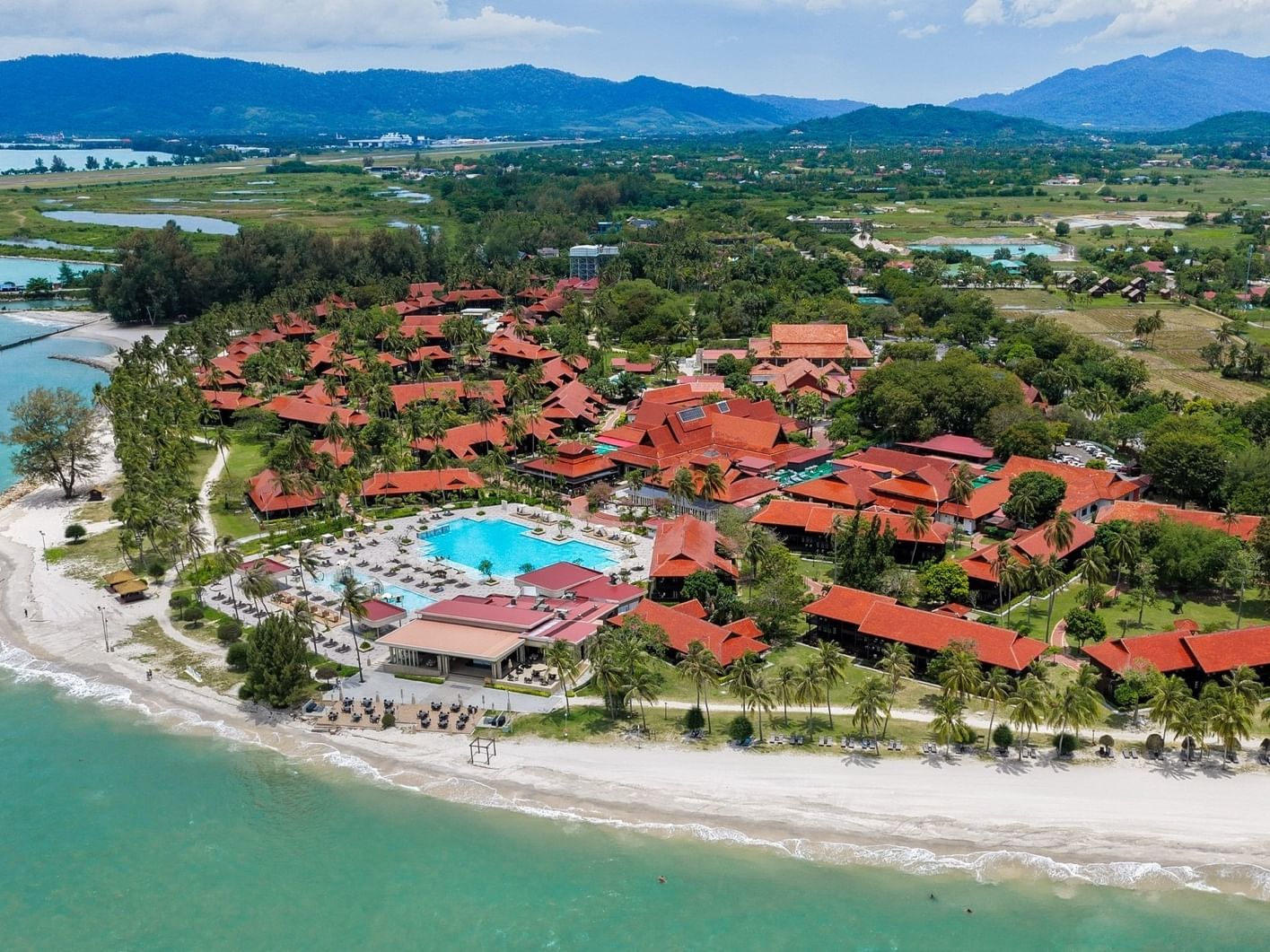 Aerial view of a resort with red-roofed buildings, a pool, beach, and distant mountains.