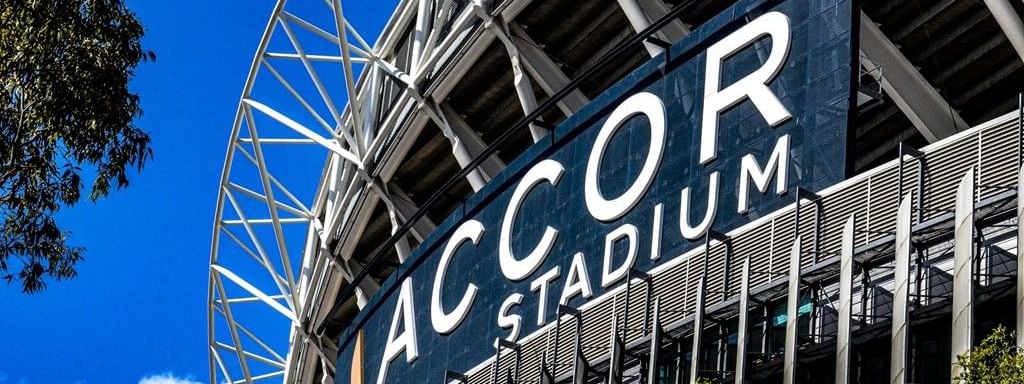 Exterior of the Accor Stadium with blue sign and surrounding trees under a clear blue sky.