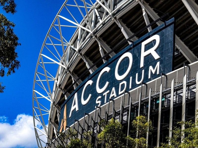 Exterior of the Accor Stadium with blue sign and surrounding trees under a clear blue sky.