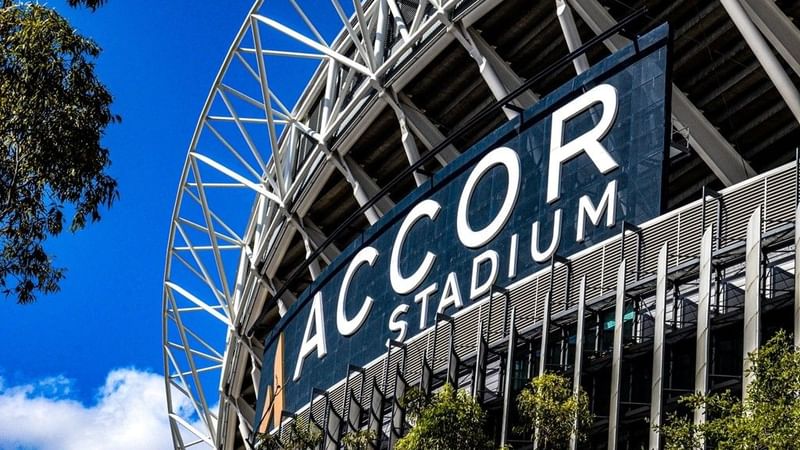 Exterior of Accor Stadium with a curved metal structure and blue signage, set against a blue sky.