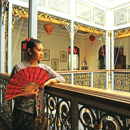 Woman styled in Nyonya Kebaya is holding a fan and striking a pose in front of the camera - Lexis Suites Penang 