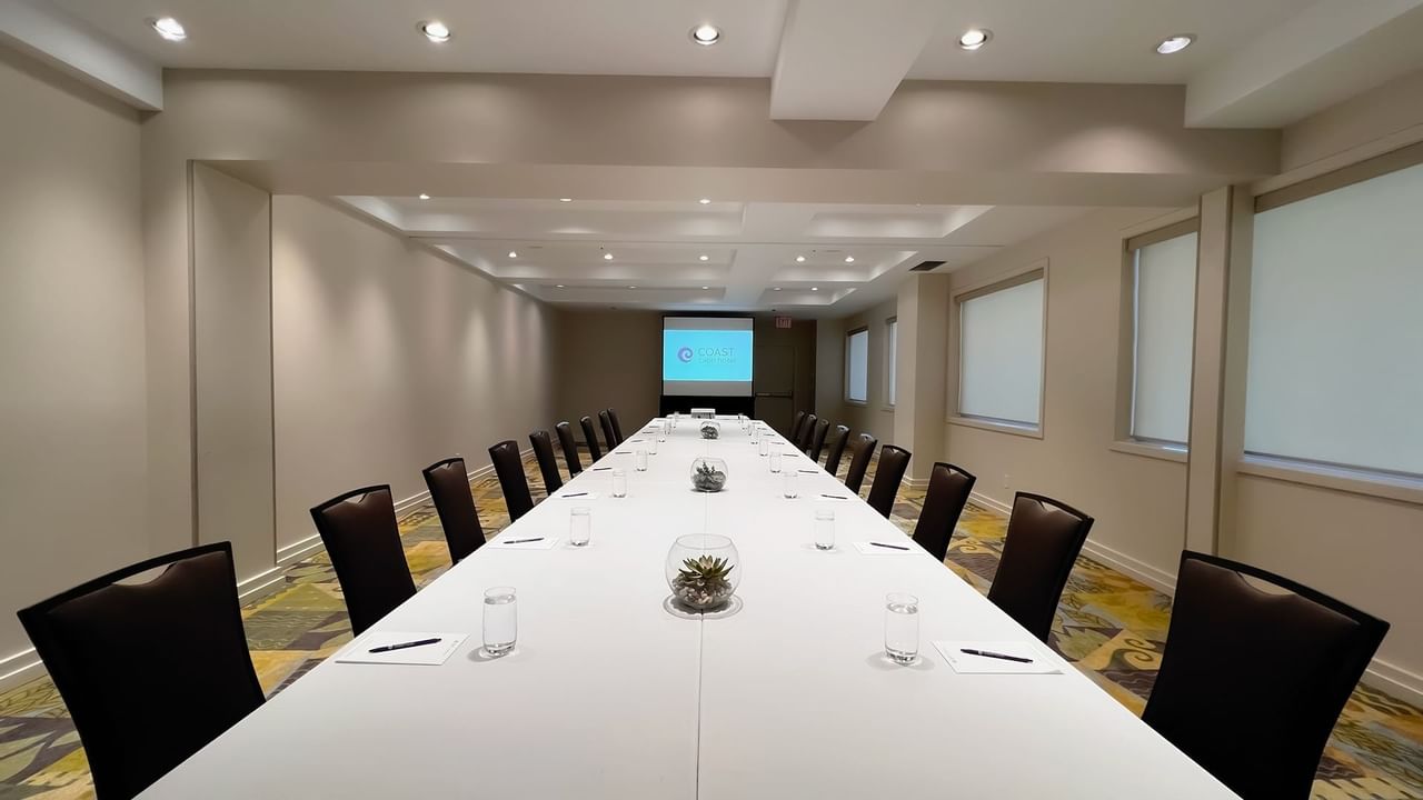Long boardroom table set with water, pens and chairs facing presentation screen
