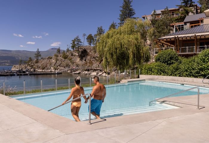 Couple exiting a pool with mountain and waterfront views, next to a modern building with green roof.