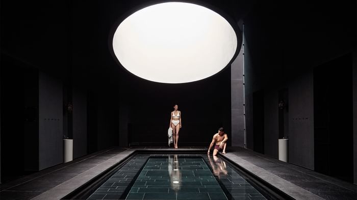 Donna e uomo accanto a una piscina in una stanza buia con un grande lucernario circolare.