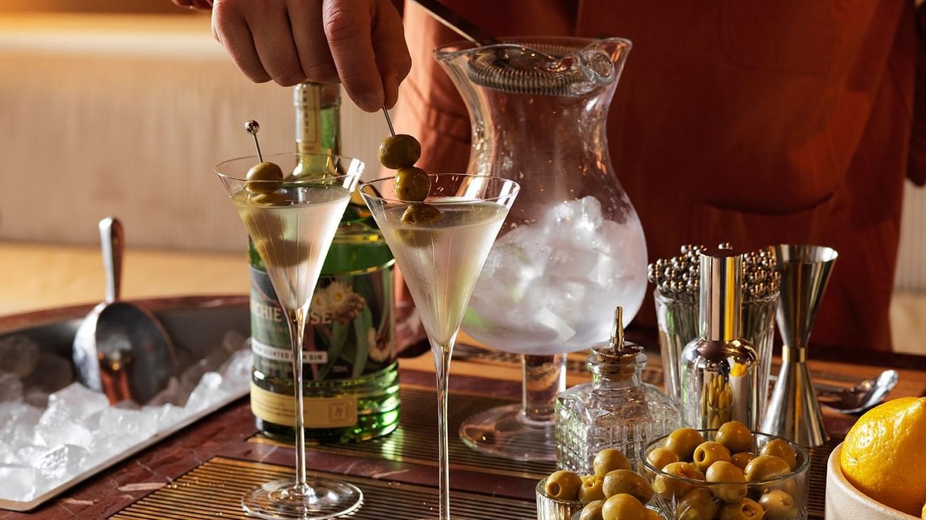 Bartender garnishing two martinis with olives in a bar setting with assorted bar tools and ingredients.