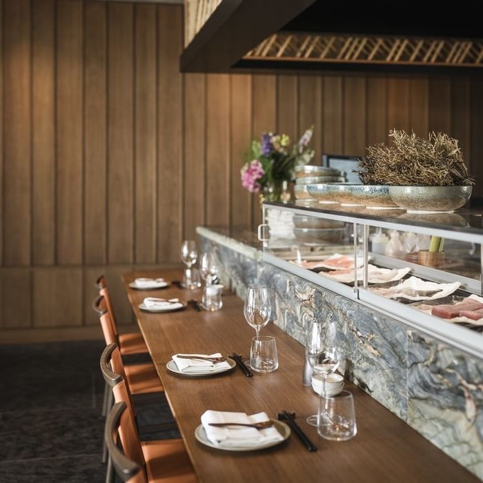 Restaurant mit gedecktem Holztisch, Weingläsern, Tellern, Stäbchen und einer Theke mit Speisen.