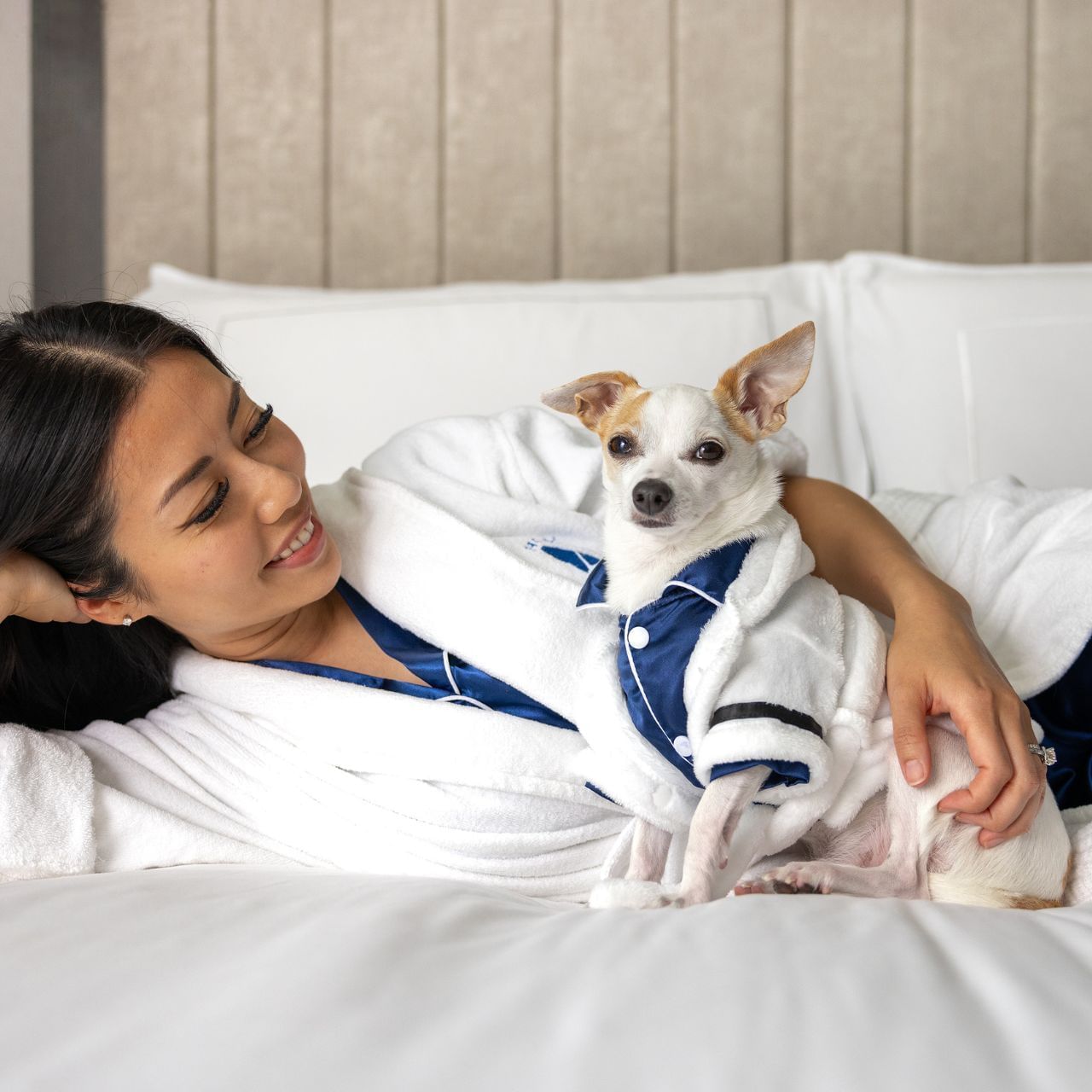 Smiling woman in white bathrobe lying on bed next to small dog wearing bathrobe and pajamas at Hotel X Toronto