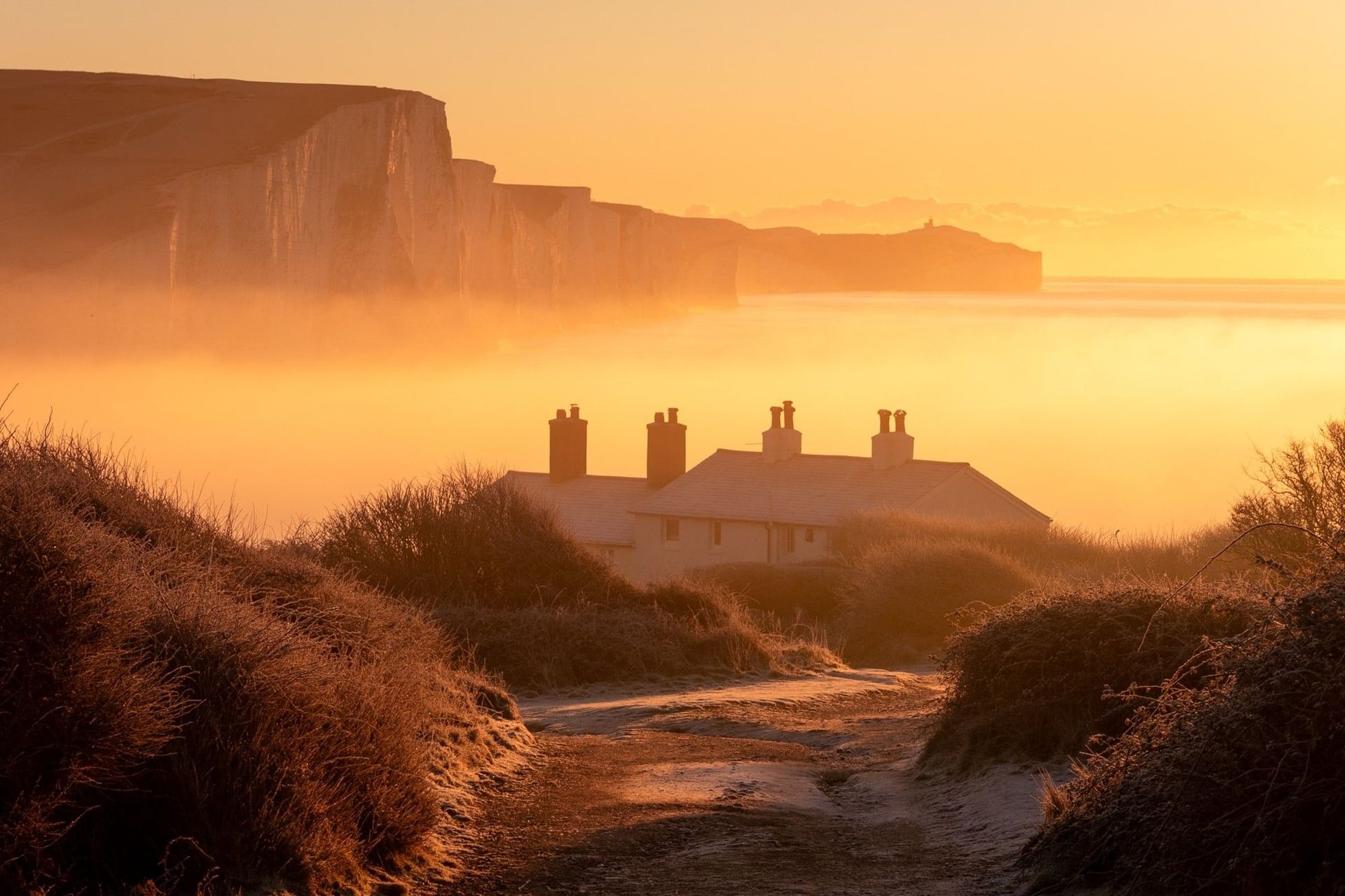 Sunset over the sea with a white house and path leading to the shore.