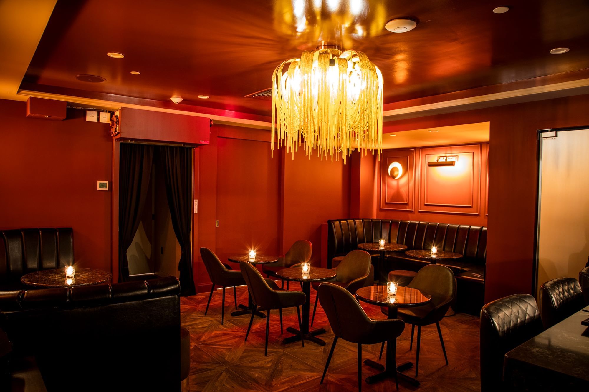 A dimly lit bar with red walls, black leather booths, and tables with candles.