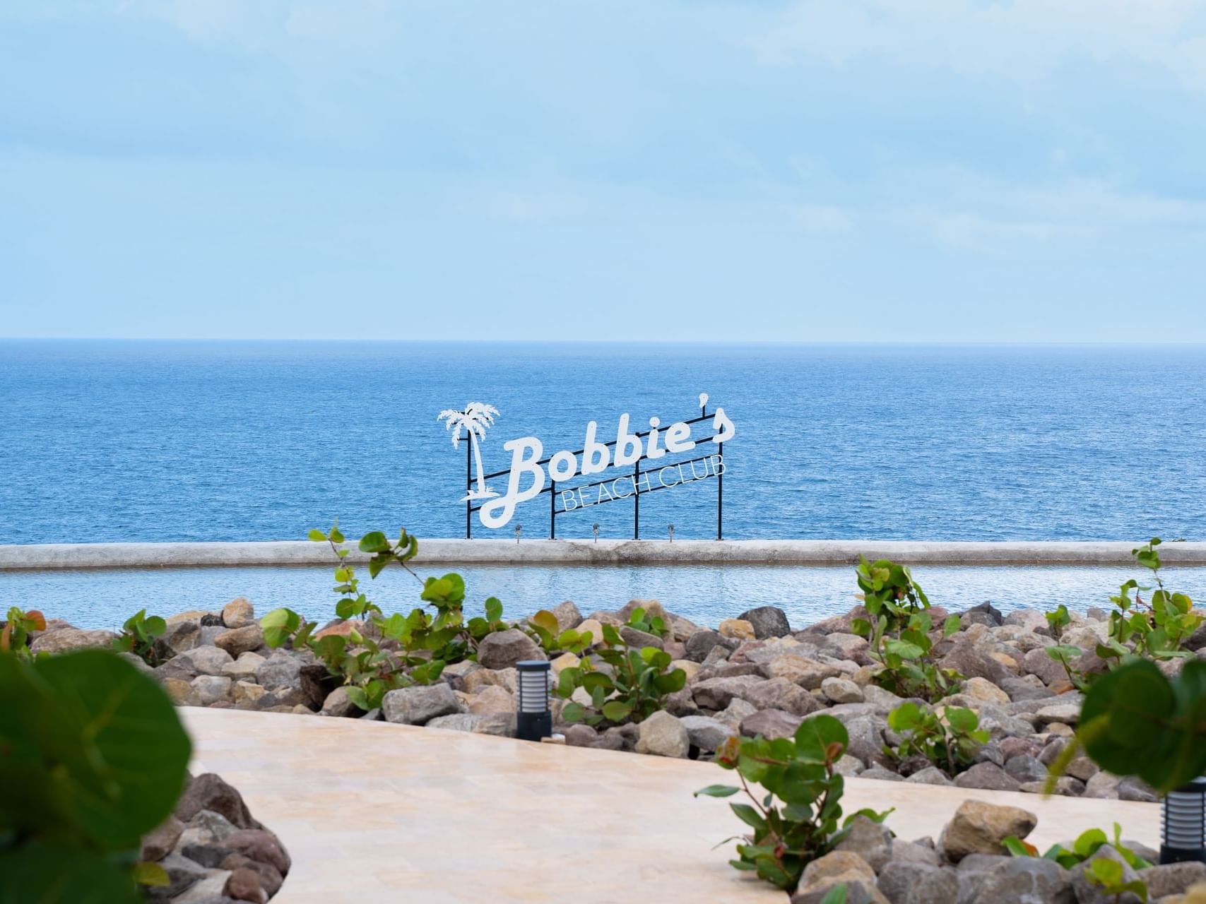 Bord van Bobbie’s Beach Club met uitzicht op een kalme oceaan, omgeven door groen en rotsachtig terrein, Golden Rock Resort