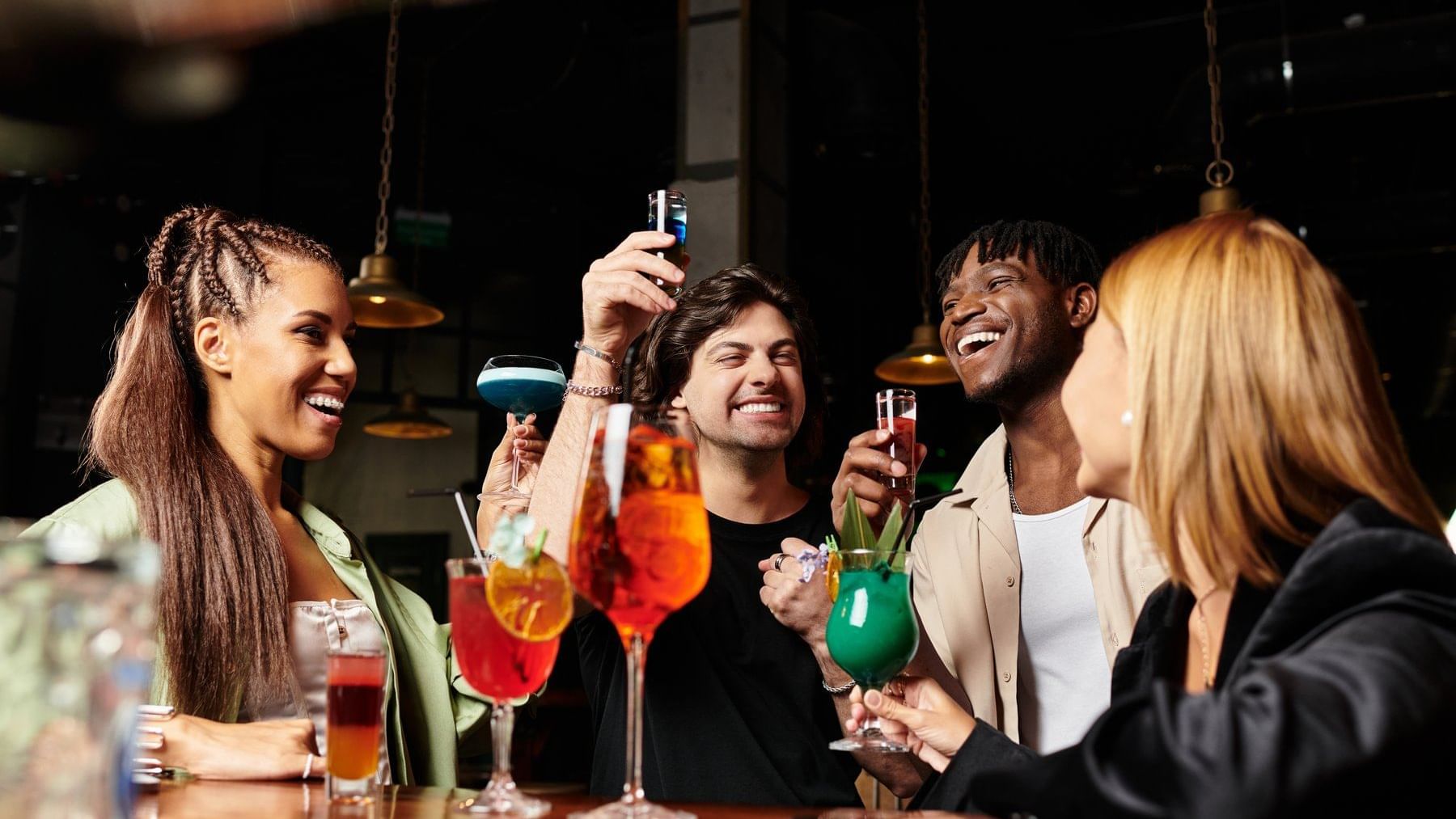 Grupo de amigos brindando felices con cócteles coloridos en el animado bar interior de Fiesta Americana Funeeq