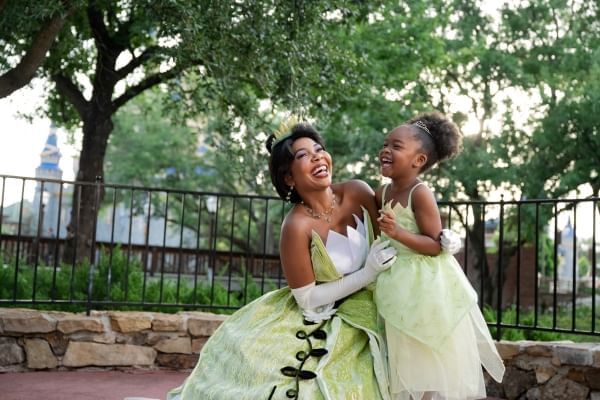 Princess Tiana in a green and white dress and crown crouched laughing beside a laughing little girl in a matching dress.