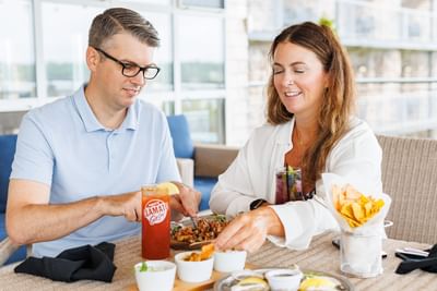 Couple dining by a table with drinks and snacks under a sunny patio area at Rodd Brudenell River Resort