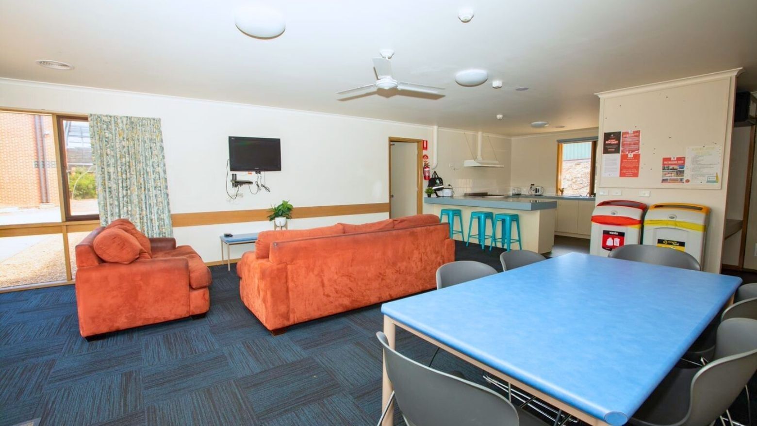 A cozy lounge with orange couches, a blue dining table, and a kitchen area at La Trobe University - Terraces.