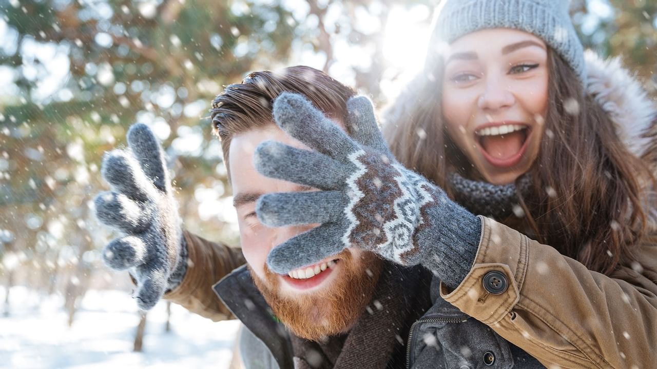 girl wearing winter gloves and hat covering mans face with snow