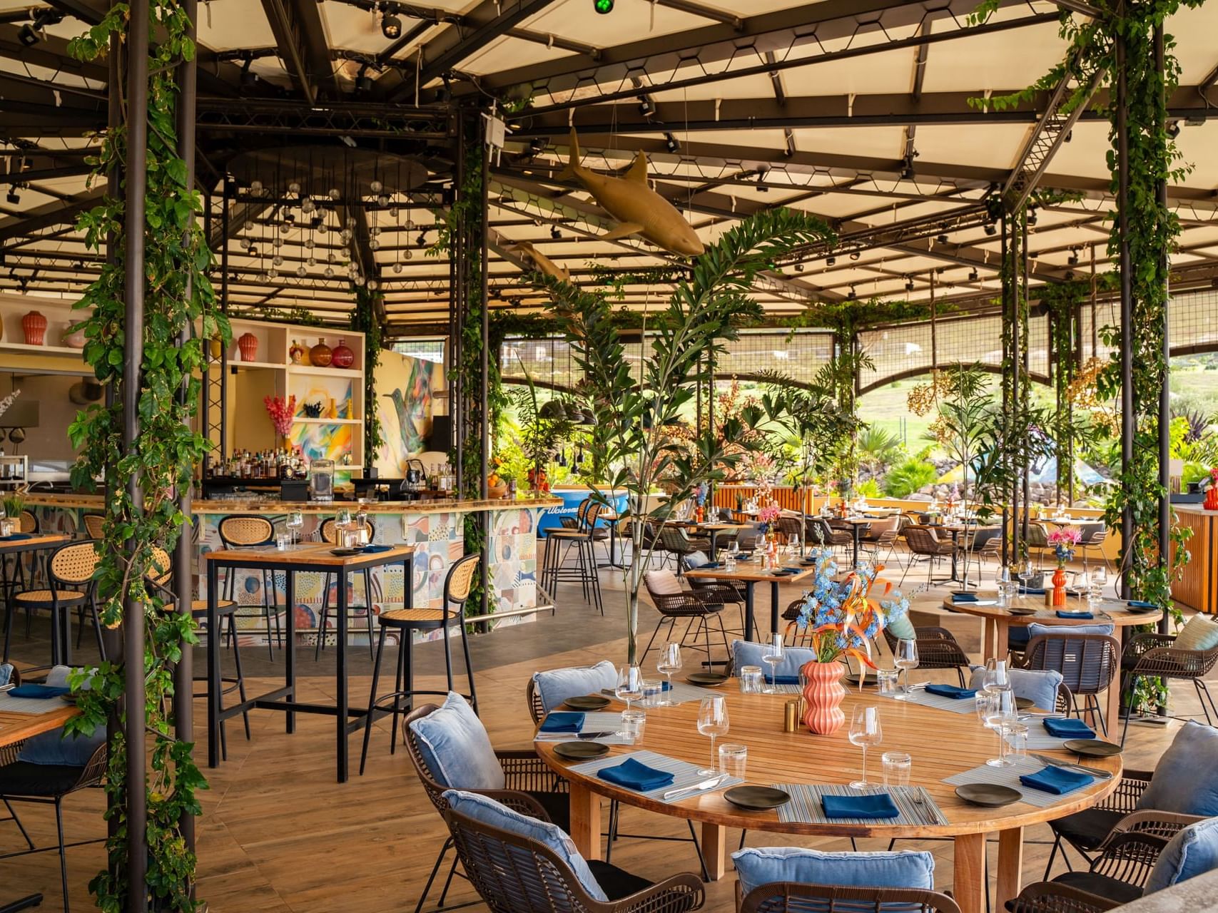 Eetgedeelte met tafels en gevlochten stoelen, gevuld met planten en decoratie in Bobbie’s Beach Club bij Golden Rock Resort