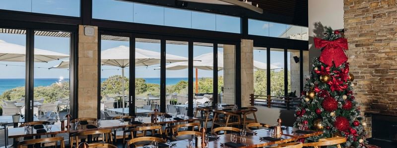 Bright restaurant with ocean views, wooden tables set for Christmas Day Lunch 2026, and a decorated tree with gifts.