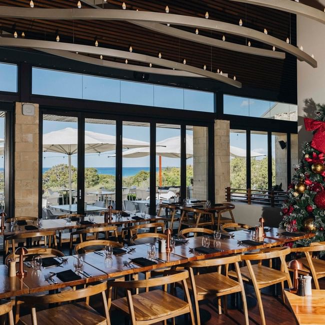 Bright restaurant with ocean views, wooden tables set for Christmas Day Lunch 2026, and a decorated tree with gifts.