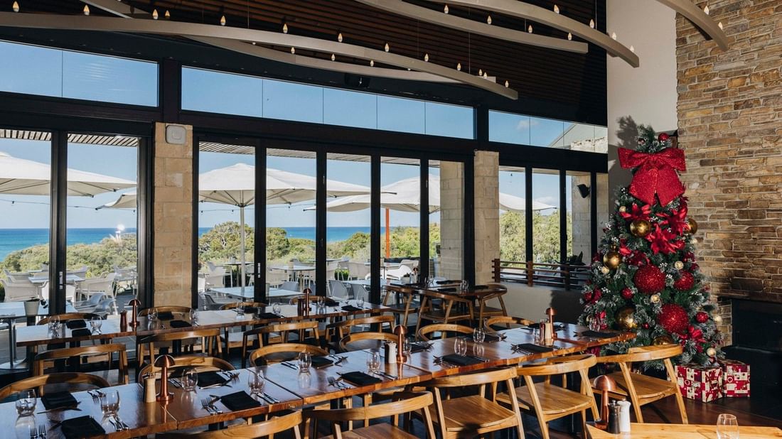 Bright restaurant with ocean views, wooden tables set for Christmas Day Lunch 2026, and a decorated tree with gifts.