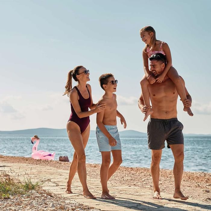 Family enjoys a sunny day at the beach with a child on a father's shoulders at Falkensteiner Punta Skala