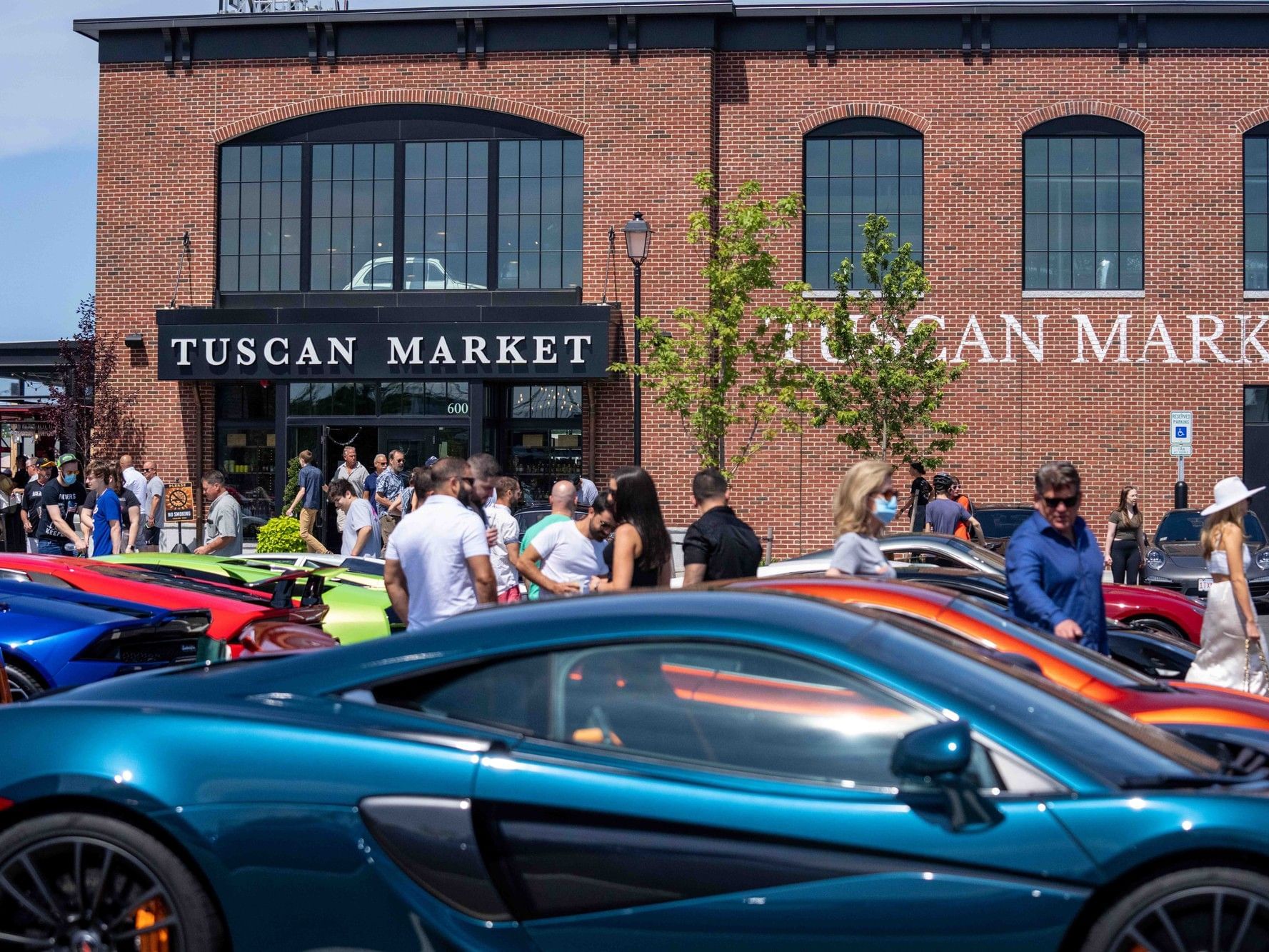 Several people gather in front of Tuscan Market, with luxury cars parked in the foreground.