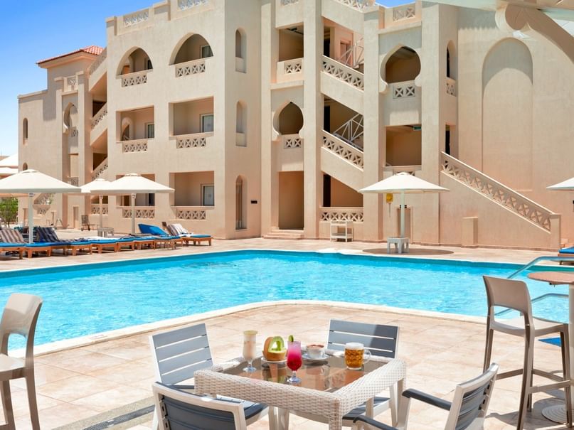 Poolbereich mit Liegen, Tischen, Stühlen und Gebäude im Hintergrund in der Sonne.