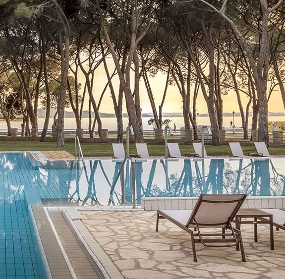 Piscina esterna circondata da alberi e sedie a sdraio, con vista sul mare al tramonto.