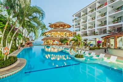 Blue pool with white lounge chairs beside white hotel building with thatched palapa structure at Playa Los Arcos Puerto Vallarta