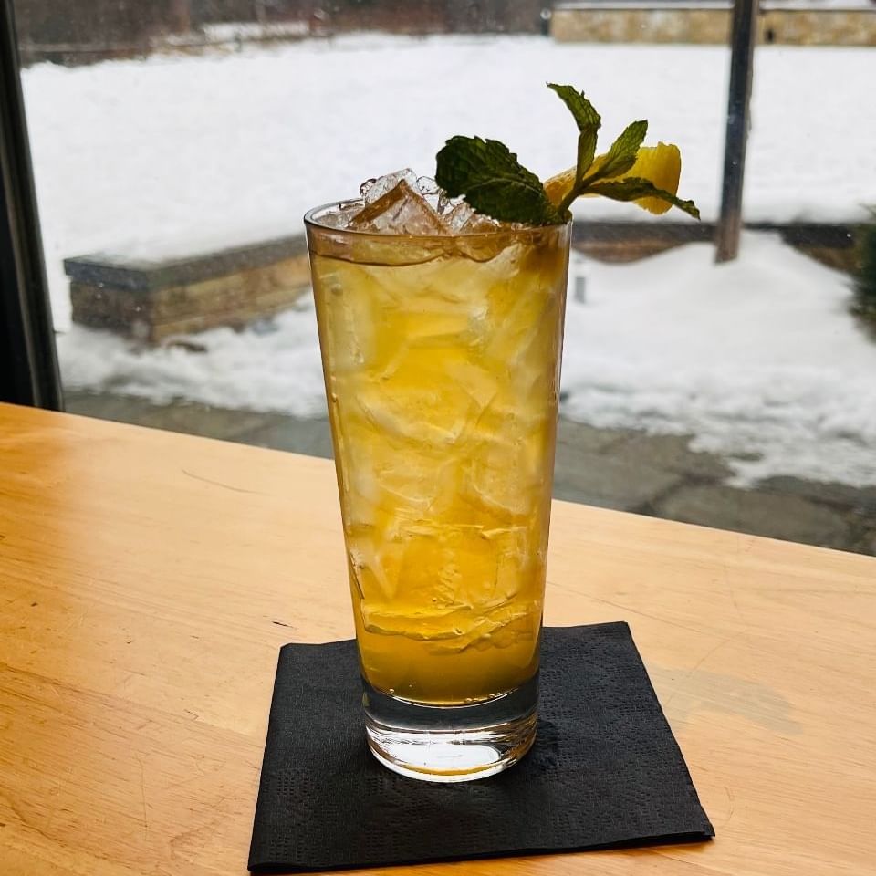 Yellow mocktail with ice and mint garnish on a black napkin against a snowy backdrop.