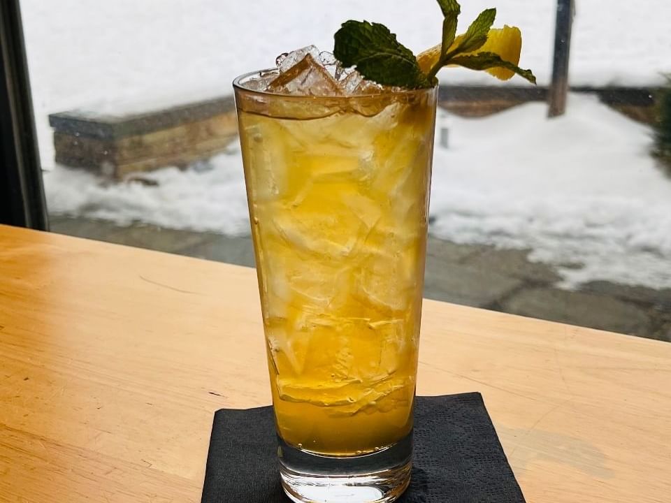 Yellow mocktail with ice and mint garnish on a black napkin against a snowy backdrop.