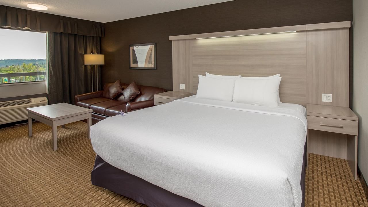 Spacious hotel room with king bed, brown leather sofa, floor lamp, and window view at Coast Calgary University Area Hotel by APA