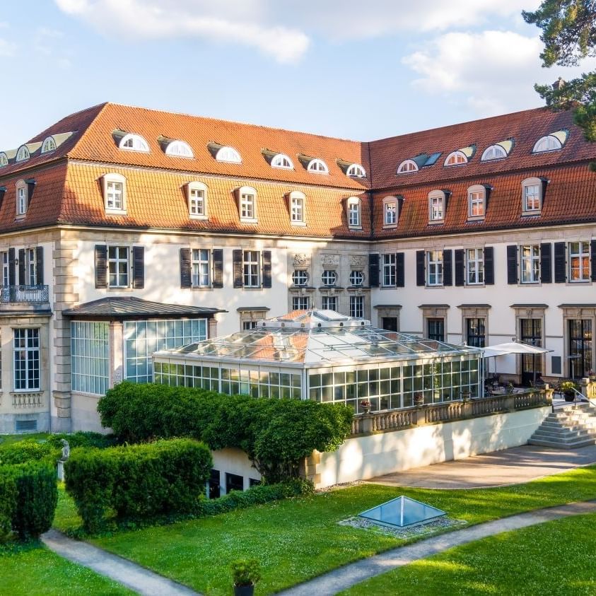Elegantes Hotelgebäude mit Glasanbau, umgeben von gepflegtem Parkgelände mit grünen Rasenflächen und hohen Bäumen.