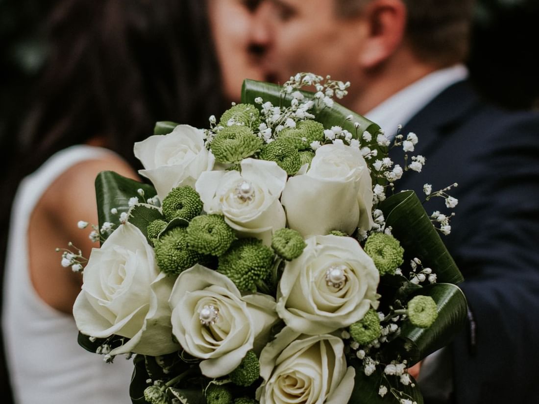 White wedding bouquet with roses in focus, with a happy wedded couple blurred in the background at The Sebel Mandurah