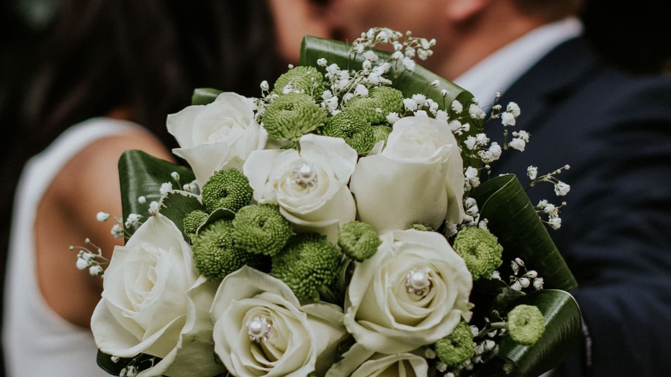 White wedding bouquet with roses in focus, with a happy wedded couple blurred in the background at The Sebel Mandurah